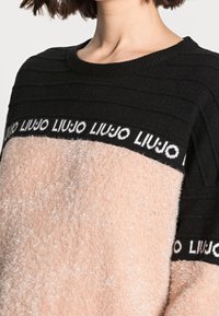 Frau trägt einen schwarzen und beigen flauschigen Pullover mit dem Markennamen "LIU JO", der in Weiß auf den schwarzen Brust- und Ärmelbändern wiederholt wird.