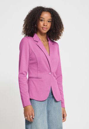 Jonge vrouw met krullend haar die een aangesloten roze blazer en lichtblauwe spijkerbroek draagt, staand tegen een effen lichte achtergrond.