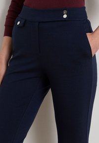 Osoba v tmavomodrých nohaviciach so striebornými gombíkmi a bočnými vreckami, s rukou odpočívajúcou v pravom vrecku, v kombinácii s bordovým topom.