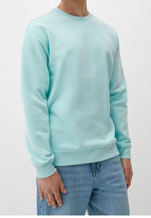 Homme portant un sweat-shirt bleu clair à col rond et un jean bleu, debout avec les mains détendues le long du corps.
