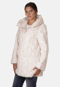 Cappotto in pelliccia sintetica bianca con superficie testurizzata, colletto oversize beige, tasche frontali e dettagli in metallo dorato. Indossato sopra una camicia chiara.