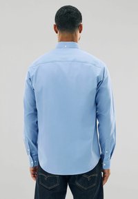 Šviesiai mėlynas ilgomis rankovėmis marškinis su standartiniu apykakle, užsegamais rankogaliais ir lygia tekstūra. Yra juostos siūlės ir subtilių siuvimo detalių.