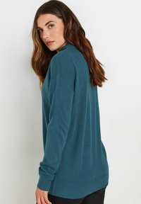 Maglione lavorato a maglia color teal con collo alto e maniche lunghe, presenta una vestibilità rilassata e un orlo a coste, visto da dietro.
