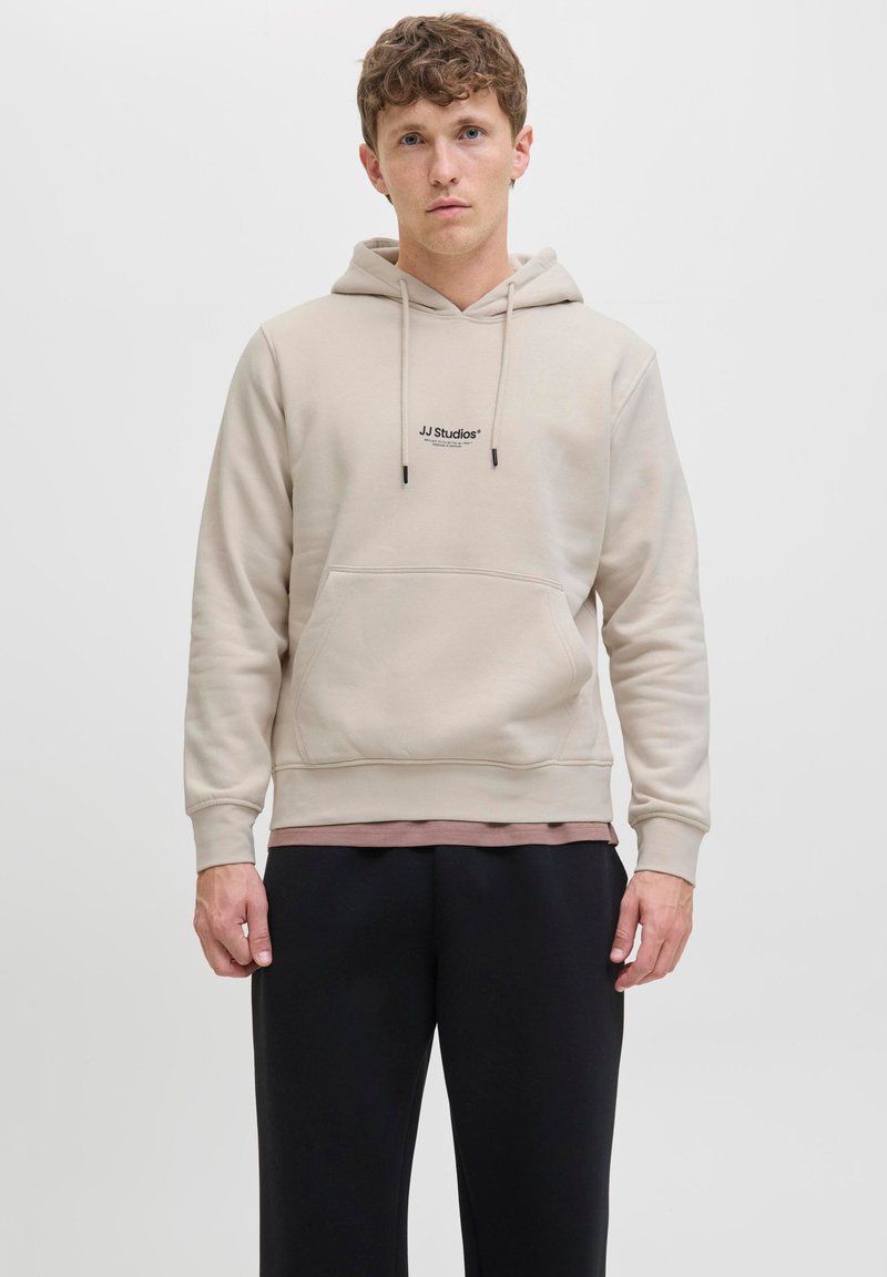 Beige Kapuzenpullover mit Fronttasche, Kordelzug und JJ Studios-Logo. Über einem geschichteten braunen Hemd getragen, kombiniert mit schwarzen Hosen.