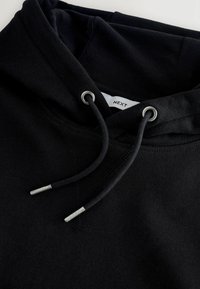 Sweat à capuche noir avec des cordons passant à travers des œillets métalliques et des embouts en métal, montrant une partie de l’étiquette intérieure indiquant "NEXT."