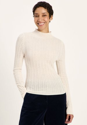 Lächelnde Frau mit kurzen lockigen Haaren, die einen weißen gerippten Langarmpullover und dunkelblaue Hosen trägt, steht mit einer Hand in der Tasche.