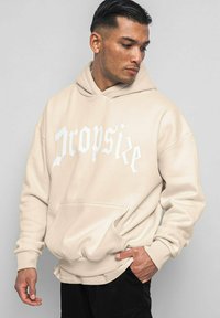 Beige Hoodie mit Fronttasche, großem weißen "Dropsize"-Logo, weichem Material, lässiger Schnitt, gerippte Bündchen und Saum. Model trägt schwarze Hosen.