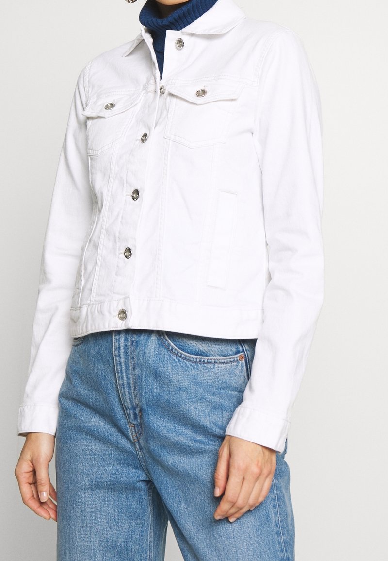 Personne portant une veste en denim blanche boutonnée par-dessus un col roulé marine et un jean en denim bleu clair, visible du cou jusqu'aux hanches.