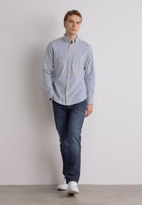 Camisa de botones a rayas en azul y blanco, combinada con jeans oscuros. El modelo lleva zapatillas blancas y está de pie contra un fondo neutro.