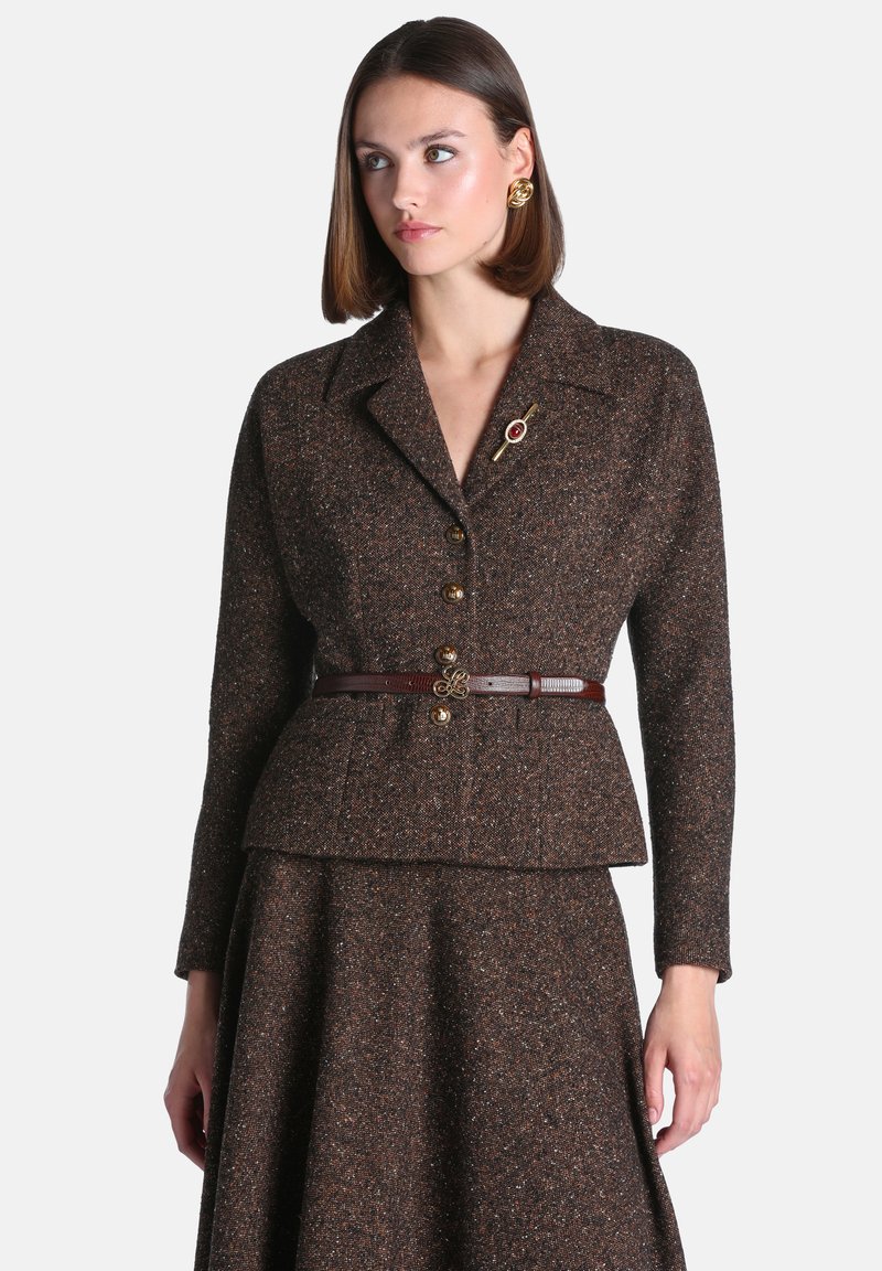 Femme portant un costume en tweed marron avec des boutons dorés, une fine ceinture marron, une broche et des boucles d'oreilles en or, regardant légèrement de côté.
