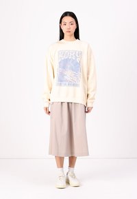 Kremfarget oversized genser med blå grafikkprint, parret med en beige midiskjørt og hvite sneakers. Behagelig, avslappet stoff.