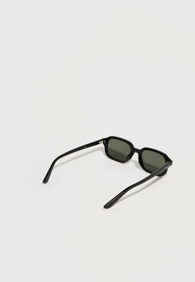 Μαύρα ορθογώνια γυαλιά ηλίου με σκοτεινούς φακούς, ανοιχτά και ακουμπισμένα σε ανοιχτό γκρι επιφάνεια.