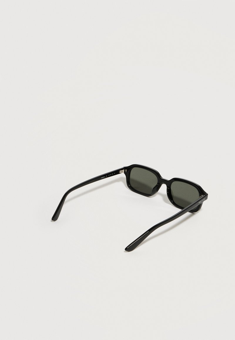 Lunettes de soleil rectangulaires noires aux verres foncés, posées ouvertes sur une surface gris clair.