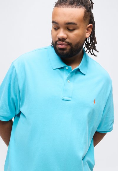 Άνδρας με μούσι και ντραντλοκς, φορώντας ανοιχτό  μπλε πόλο πουκάμισο, κοιτάζοντας προς τα κάτω και πλάγια, με απλό φόντο.