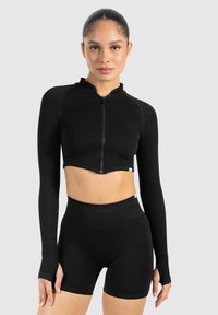 Sort cropped langærmet top med høj krave og lynlås foran, parret med højtaljede sorte shorts. Glat tekstur, tætsiddende design.