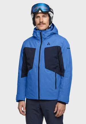 Man wearing blue and black ski jacket, white helmet, and blue ski goggles standing against a plain background.