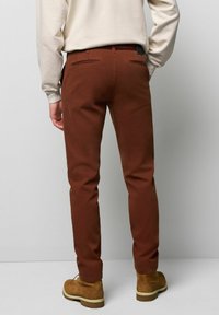 Bruine katoenen broek met een slim fit, voorzien van achterzakken en een gevlochten riem. Gepaard met lichtgekleurde laarzen.