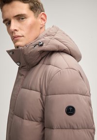 Giubbotto impermeabile marrone chiaro, con cappuccio, dettagli di cucitura e una patch rotonda nera sul maniche sinistro. Bottoni a pressione argento visibili.