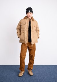 Braune Puffjacke mit einem mit Fell gefütterten Kragen, mehreren Fronttaschen, kombiniert mit braunen Cordhosen und beigen Schuhen. Schwarze Mütze auf dem Kopf.