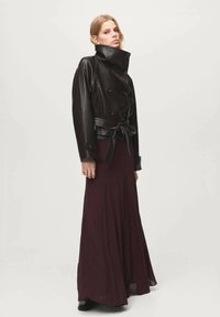 Czarna skórzana kurtka dwurzędowa z wysokim kołnierzem i paskiem, zestawiona z bordową spódnicą do ziemi. Gładkie tekstury i smukła sylwetka.