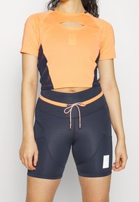 Crop top sportivo arancione con finitura texturizzata; presenta una apertura a forma di chiave e accenti blu navy. Pantaloni corti blu navy a vita alta con coulisse.