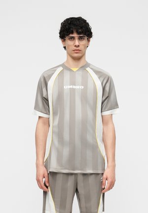 Νέος άνδρας με σγουρά μαλλιά και γυαλιά φοράει αθλητική φόρμα γκρι με ρίγες και λευκές και κίτρινες λεπτομέρειες, στέκεται μπροστά σε απλό ανοιχτόχρωμο φόντο.