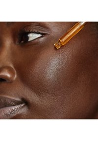 Un contagocce rilascia un liquido ambrato sulla guancia di una donna dalla pelle scura vicino all'occhio, evidenziando l'applicazione di prodotti per la cura della pelle e la texture del viso.