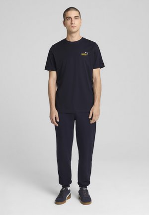 Marineblauwe katoenen t-shirt met korte mouwen en een klein geel Puma-logo op de borst, gecombineerd met bijpassende joggers en donkere schoenen.