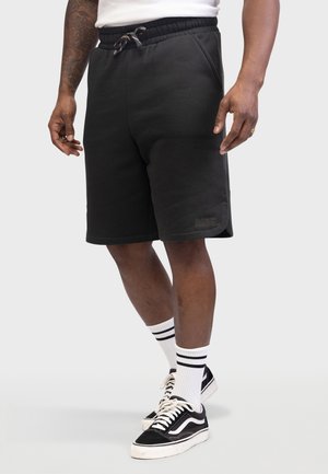 Shorts de sport noirs avec une taille élastique et un cordon de serrage, dotés de poches latérales et d'un petit logo "REBEL" en gris. Portés avec des baskets noires et des chaussettes rayées.