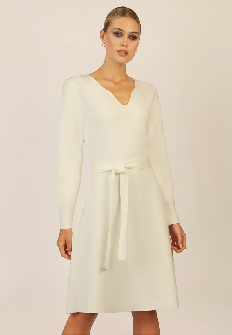 Weißes Kleid mit V-Ausschnitt und langen Ärmeln, figurbetontem Oberteil, weit schwingendem Rock und einem Stoffgürtel, der in der Taille gebunden wird. Weiche Textur, minimalistisches Design.