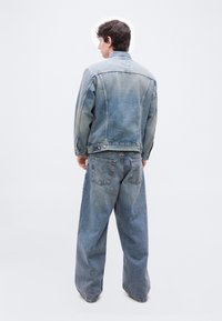 Άνδρας που φοράει ξεθωριασμένο ανοιχτό μπλε τζιν μπουφάν και φαρδιά τζιν, στέκεται με την πλάτη προς την κάμερα σε λευκό φόντο.