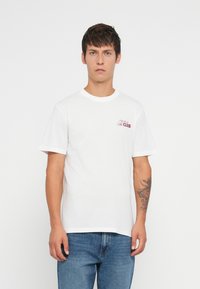 Hvit bomull t-skjorte med korte ermer, rund hals, med et lite rødt grafisk tekstdesign på venstre bryst. Brukt sammen med blå jeans.