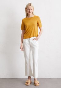 Mosterdkleurig T-shirt met korte mouwen, gecombineerd met witte, enkellange jeans. De outfit heeft textuur-sandalen en een relaxed fit met een afgewerkte rand.