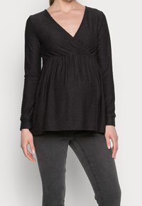 Černý těhotenský top s dlouh ým rukávem a výstřihem do V, se strukturou bodových vzorů a nařaseným empírovým pasem, doplněný tmavě šedými džíny.
