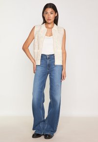 Gilet en tricot sans manches de couleur beige clair avec un col, associé à un jean bleu taille haute à jambes larges. Les deux pièces ont une coupe décontractée.