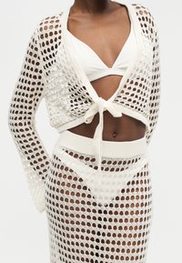 Personne à la peau foncée portant un cardigan blanc en crochet noué à la taille sur une jupe en crochet assortie et un haut et bas de bikini blancs.