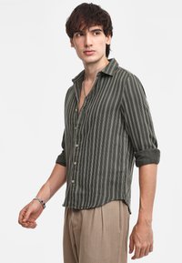 Camicia button-up verde scuro con righe verticali bianche, maniche arrotolate e vestibilità rilassata. Presenta bottoni in legno chiaro e una consistenza morbida.