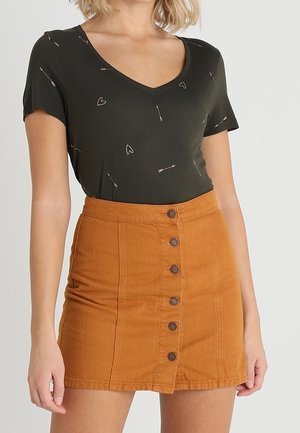 Kobieta nosi czarną bluzkę z krótkim rękawem i dekoltem w serek, zdobioną małymi nadrukami serc i strzał, w połączeniu z brązową mini spódnicą z wysokim stanem i guzikami.