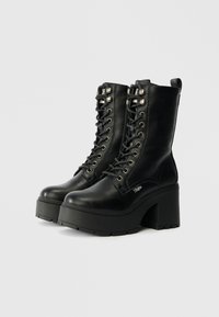 Μαύρες δερμάτινες μποτές με κορδόνια στον αστράγαλο, με χοντρή σόλα και ψηλό τετράγωνο τακούνι, διαθέτοντας μεταλλικά θηλάκια και λογότυπα.