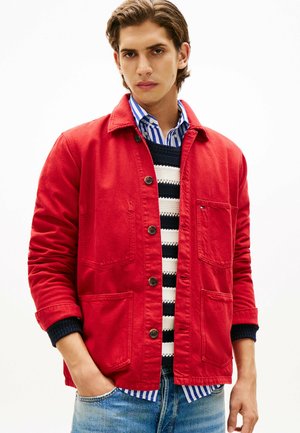 Ung mand iført en rød knappet jakke over en sort og hvid stribet sweater og en blå stribet skjorte, med hænderne delvist i jeanslommerne.