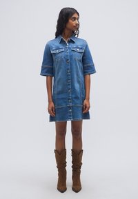 Φόρεμα από τζιν πουκάμισο, με κοντά μανίκια, ανοιχτό μπλε χρώμα, εμπρός με κουμπιά, δύο τσέπες στο στήθος, μέχρι το γόνατο. Φορεμένο με καφέ μποτάκια αστραγάλου.