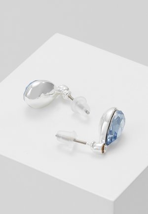 Boucles d'oreilles clous en argent avec des pierres précieuses en forme de goutte d'eau bleu et des attaches en plastique transparent posées sur une surface blanche.