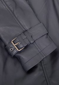 Detail eines dunklen Lederbekleidungsstücks, das einen Gürtel mit einer silbernen Metallschnalle zeigt. Glatte Textur mit sichtbaren Nähten um den Gürtel.