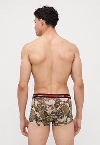 Boxers pour hommes avec un motif floral beige, dotés d'une ceinture noire avec le logo rouge "HUGO", fabriqués en tissu doux et texturé.