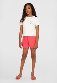Kortærmet hvid t-shirt med et palmetryk på brystet, parret med lyserøde, plisserede shorts. Model iført lyserøde flip-flops.