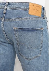 Jasnoniebieskie dżinsy z denimu z skórzaną metką na pasku. Posiadają szwy tylnej kieszeni w zakrzywionym wzorze i lekko postrzępioną teksturę.