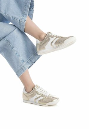 Zapatillas beige y blancas con detalles dorados texturizados, que presentan un perfil bajo, punta redonda y suela de goma, llevadas con pantalones de denim claros.