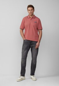 Man in een stofrozenkleurig poloshirt met autologo, donkere versleten jeans en witte sneakers, staand met één hand in zijn broekzak.