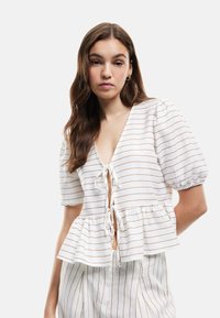 Top peplum in tessuto leggero con strisce bianche e marroni, scollatura a V profonda, chiusura a fiocco e maniche a sbuffo. Progettato per una vestibilità rilassata.