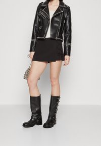 Veste de biker en cuir noir avec des clous, mini-jupe noire ajustée avec des détails de lacets sur les côtés, et bottes noires jusqu'aux genoux avec des accents métalliques.